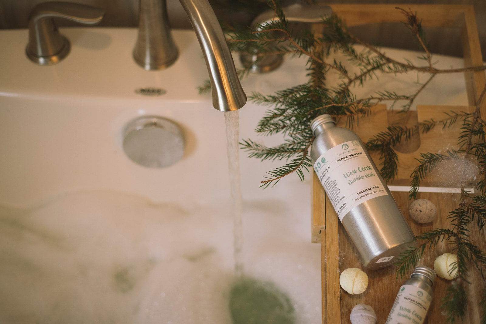 Water pouring into a bubble bath. Evergreen branches and Nezza Naturals bath products are in view on a bath tray in a softly lit room.