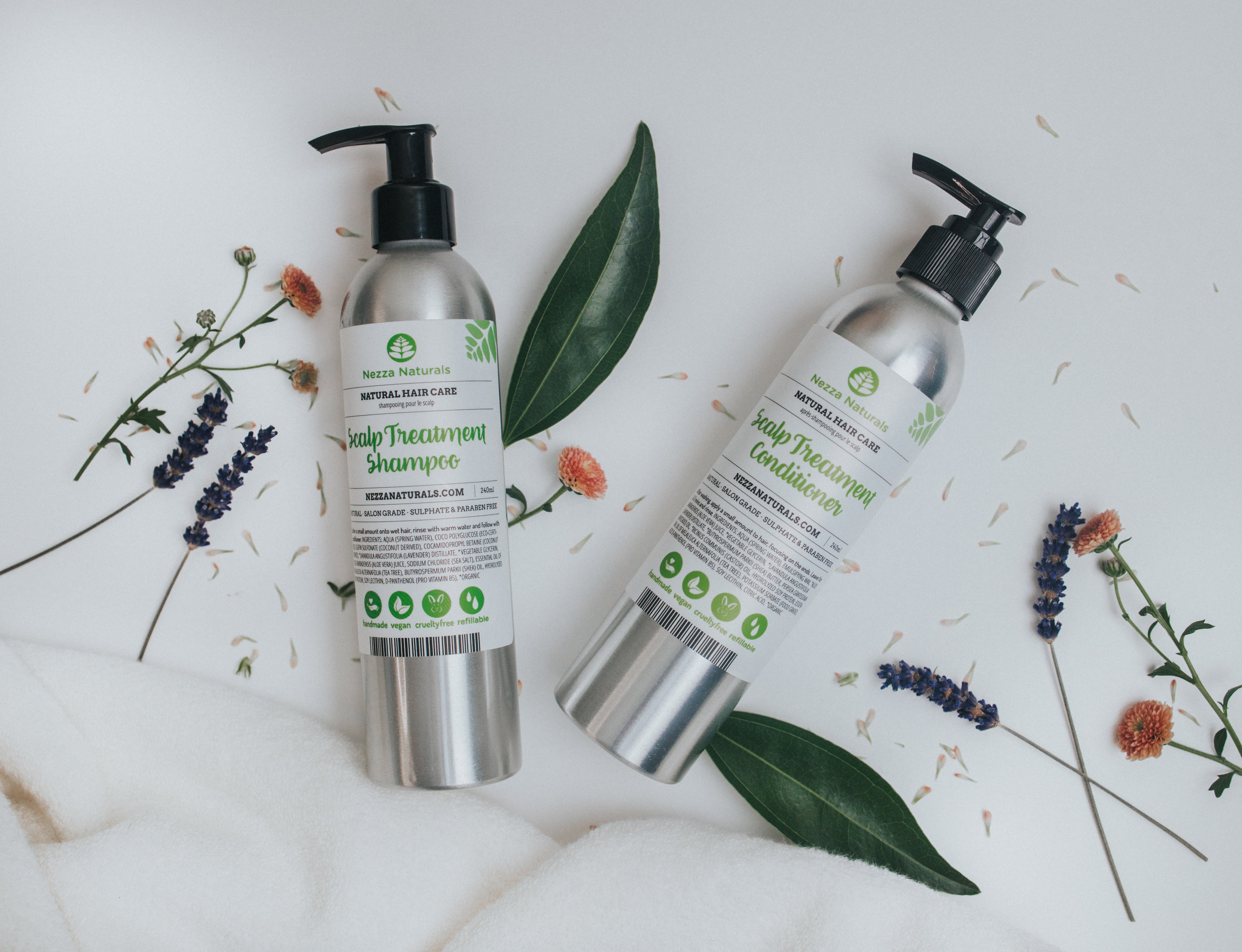 Nezza Naturals Shampoo and conditioner in metal bottles laying on a white surface with scattered leaves and flowers.